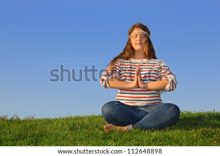 Fat girl in jeans sits at green grass and meditates at background of blue sky.