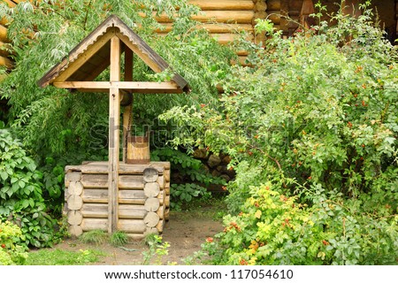 Log well with bucket and wooden carved decorations stands among green bushes.