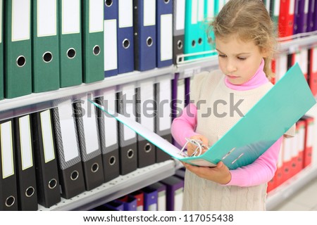 Serious looks at big green folder and stands near to shelves with folders.