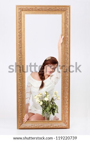 Beautiful woman in white openwork dress sits on floor behind big frame near vase with dry roses.