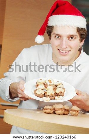 Young smiling confectioner in santa hat shows plate with cookies and cakes on it