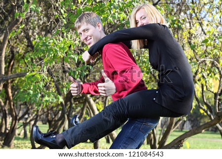 Happy woman sits on back of man and man thumbs up in park at sunny autumn day.