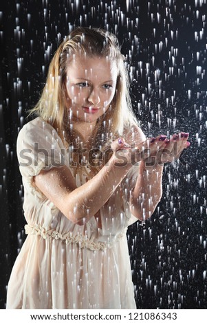 Beautiful girl in transparent blouse stands in rain in black studio and catches drops by palms.