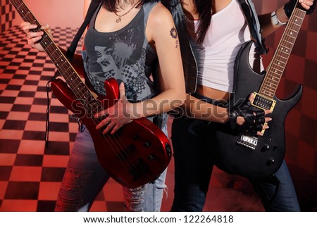 Part of bodies of two women playing electric guitar in studio with checkered background in red light and smoke.