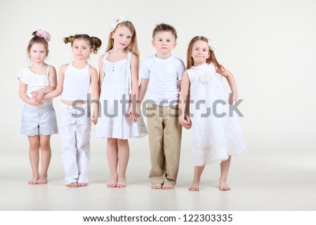Four cute little girls and one boy in white clothes stands and hold hands.