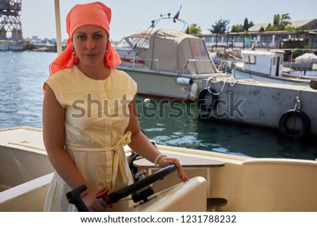 Woman in light dress and red head shawl at steering wheel on boat.
