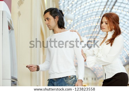 Dark-haired man with credit card and red-haired woman stand near to cash dispenser