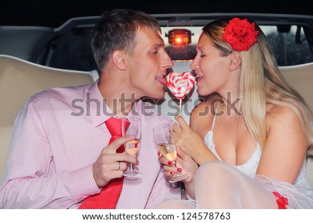 Young bride and groom sits in car, hold glass of champagne and lick one candy heart. Pink wedding.