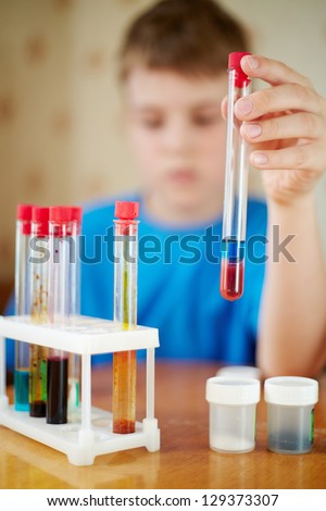 Boy in blue t-shirt sits at table with chemical reagents and holds test tube in his hand, focus at fingers and tube