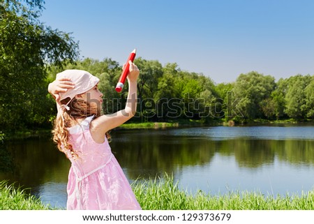 Girl in pink dress and panama from sun stands on bank of pond and writes with large red pencil on sky