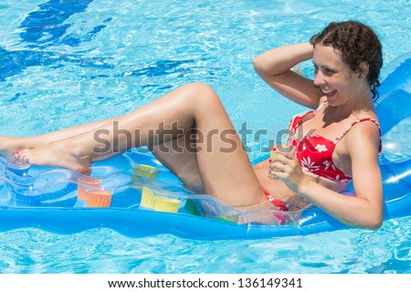 Smiling woman swimming in the pool on an inflatable mattress with a drink in hand