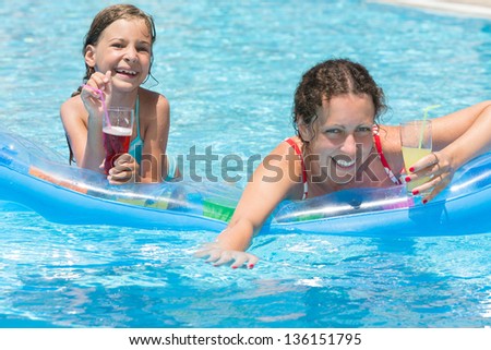 Happy and smiling mother and daughter swimming in the pool on an inflatable mattress with a drink in hand