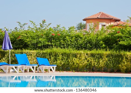 Poolside with deck-chairs and folded umbrella, bushes with flowers and villa