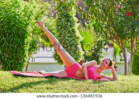 Young woman does leg swing lying on matting in morning garden