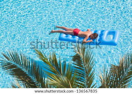 A boy floats on an inflatable mattress in the pool face down near palm leaf.