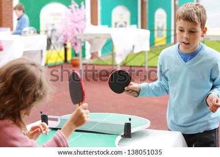 Little girl and boy in blue play table tennis in park at summer day. Focus on boy.