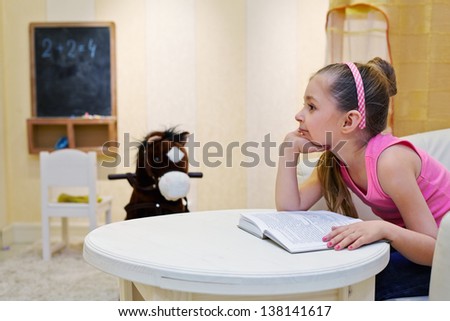Little girl dreams propping chin sitting in big armchair and viewing book in playroom