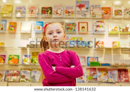 Little girl stands with arms folded on chest in book department of store against showcase with books