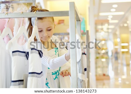 Little girl looks with interest upon dresses hanging on rack in clothing store