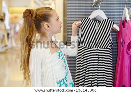 Little girl takes hanger with striped gown from stand in clothing store