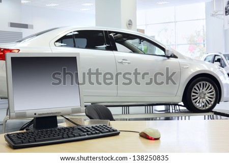 Computer stands on table and new white car stands in office of shop selling cars.