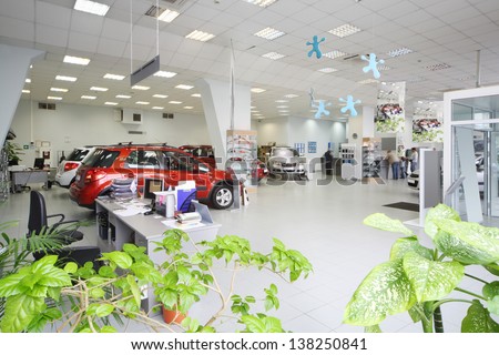 New shine cars stand in big office of shop selling cars near tables with computers.