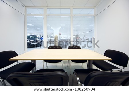 Chair and table in small room and new cars in office of car shop through window visible.