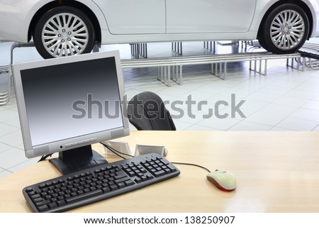 Computer stands on wooden desk and new car stands in office of car shop.