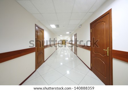 Long light hallway with brown wooden doors and white shiny floor.