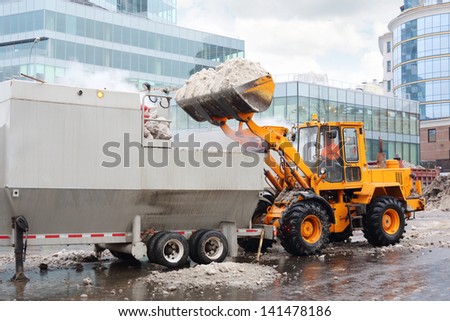 Bulldozer loads snow to truck for snow melting on street in city at cloudy day.