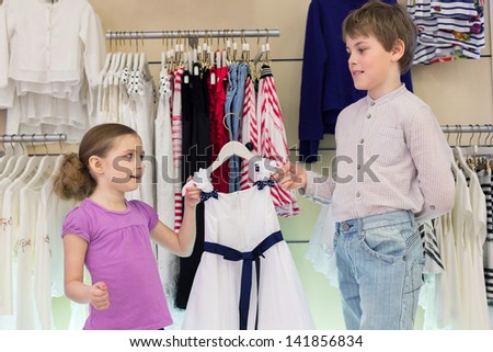 The brother helps sister to choose clothes in shop of children clothing