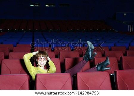 Happy beautiful woman in red glasses lifted her legs on chair in movie theater.
