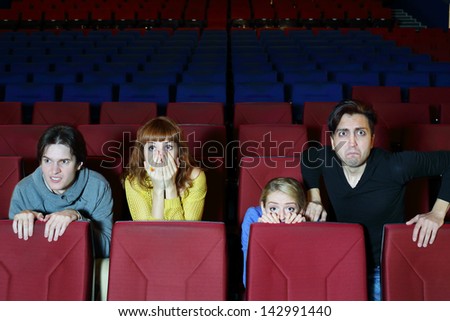 Four young scared friends see movie in cinema theater. Girl hides behind chair.