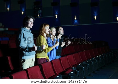 Four happy friends stand, look at screen and cheer in cinema theater. Focus on left pair.