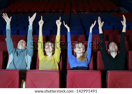 Four happy young friends sit in cinema theater and pull hands up.
