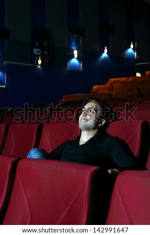 Young happy man watches movie and rests in cinema theater with red seats.