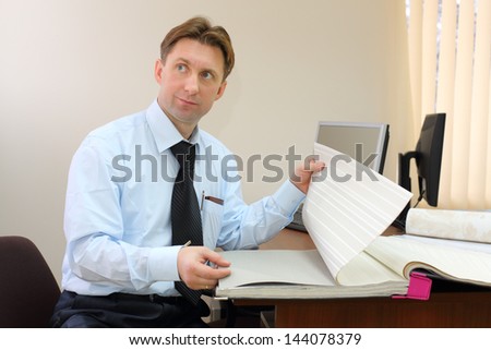 Man wearing tie sits in office of realtor, looks away and scrolls samples of wallpaper.