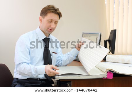 Man wearing tie sits in office of realtor and looks samples of wallpaper.