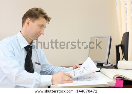 Man wearing tie sits in office of realtor and looks drawings of apartment.
