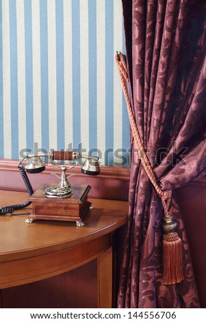 Vintage phone on table near wall with striped wallpaper and curtain with cord brush.