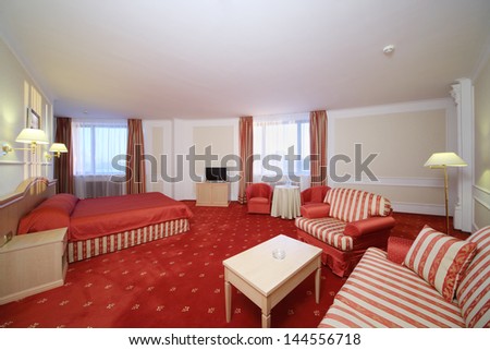 Simple room with double bed with red linen, red carpet and armchairs.