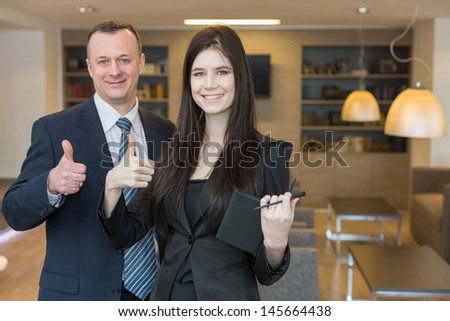 Smiling man and woman in business suits standing and doing a thumbs up, focus on a girl.
