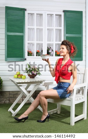 Happy woman in shorts sits at table next country house and holds plate with strawberries.