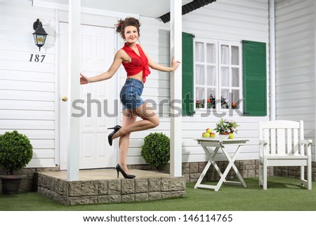 Smiling woman in shorts poses next to simple entrance door of country house.