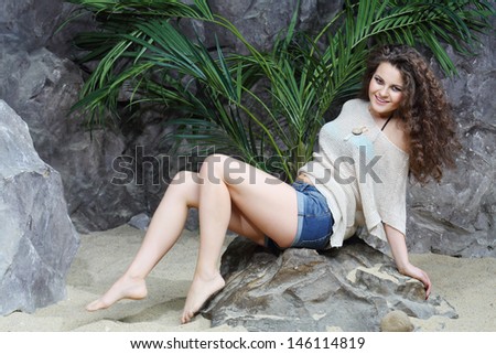 Beautiful woman in shorts and blouse sits on stone next to gray rocks with palm tree.