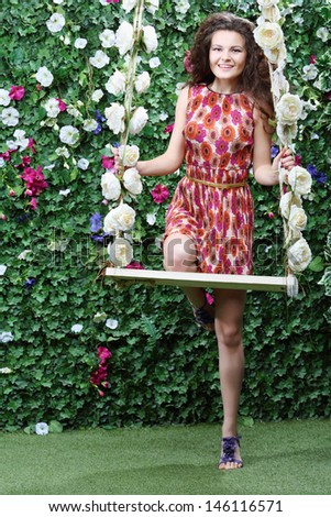 Smiling woman stands next swing overgrown with flowers next to green hedge with flowers.