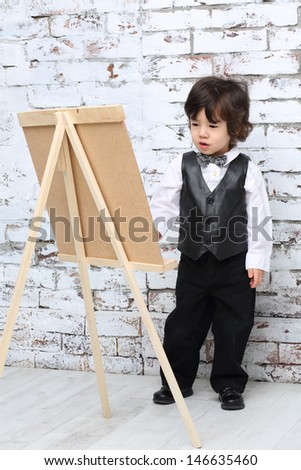 Little boy in bow tie stands next to easel in studio with white brick wall.