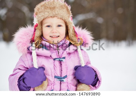 Half-length portrait of smiling little girl in pinky jacket with fur collar and earflapped hat in winter park