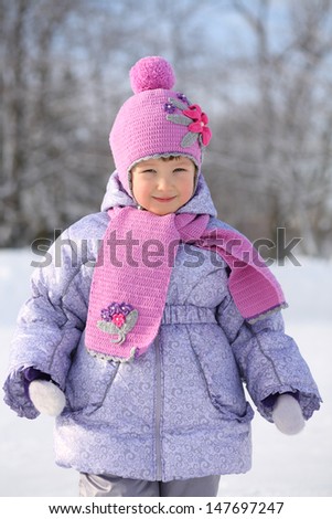Happy little girl wearing in pink scarf and hat smiles next winter forest.