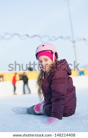 Smiling little girl in a helmet and knee pads sitting at the rink in winter